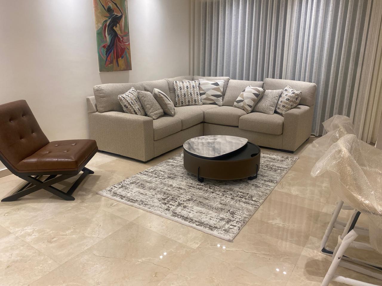 L-shaped beige sectional with patterned cushions, round marble-top coffee table, brown leather lounge chair, and sheer curtains in a modern living room.