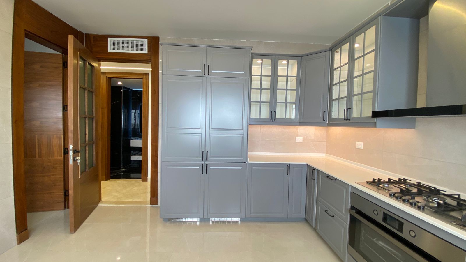 644375b1-4547-4a7b-b133-1bcc496ada08 Modern grey kitchen with glass-front upper cabinets, beige backsplash, and stainless steel oven and gas cooktop.