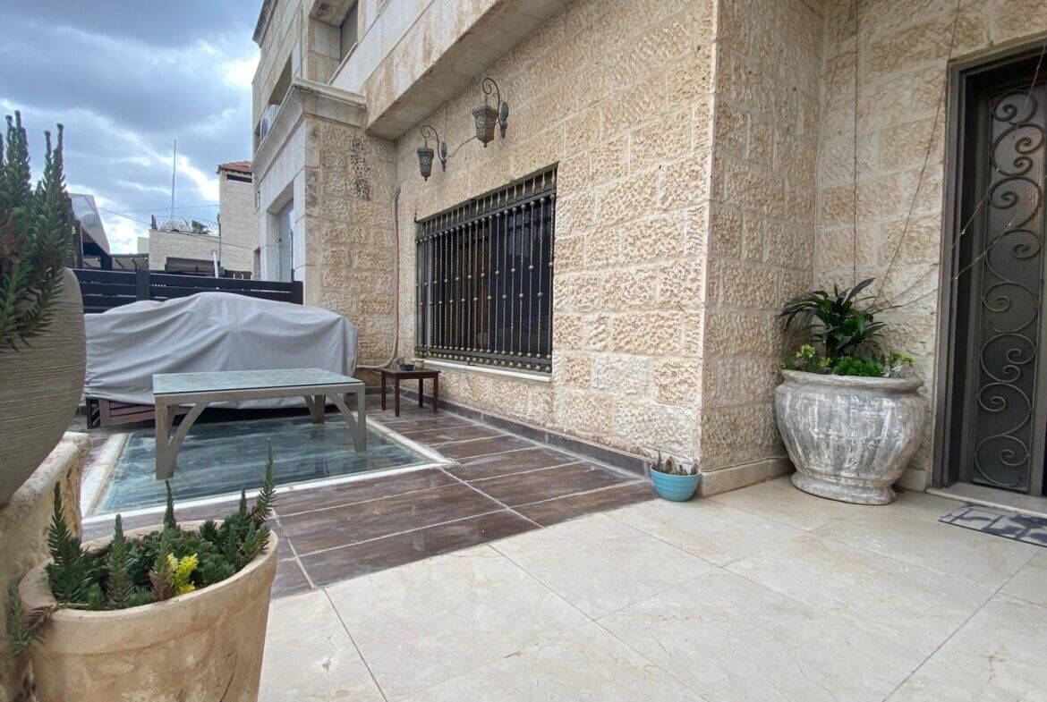 Stone-walled courtyard with a covered outdoor table beside a small glass pool and potted plants along the wall edge.