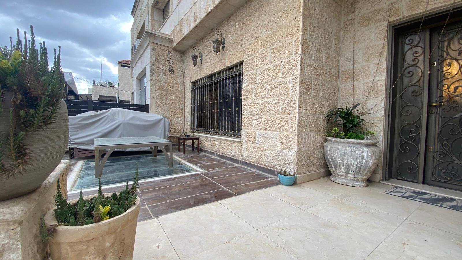 6592ae08-0149-498b-83fb-b1eef459320a Stone-walled courtyard with a covered outdoor table beside a small glass pool and potted plants along the wall edge.