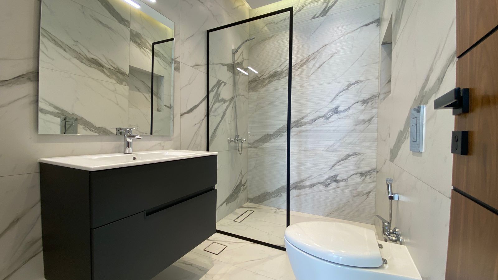 66229c3a-6238-4120-a46f-90dc8c14306d Modern bathroom with white marble walls, a black floating vanity, and a glass-framed shower area.