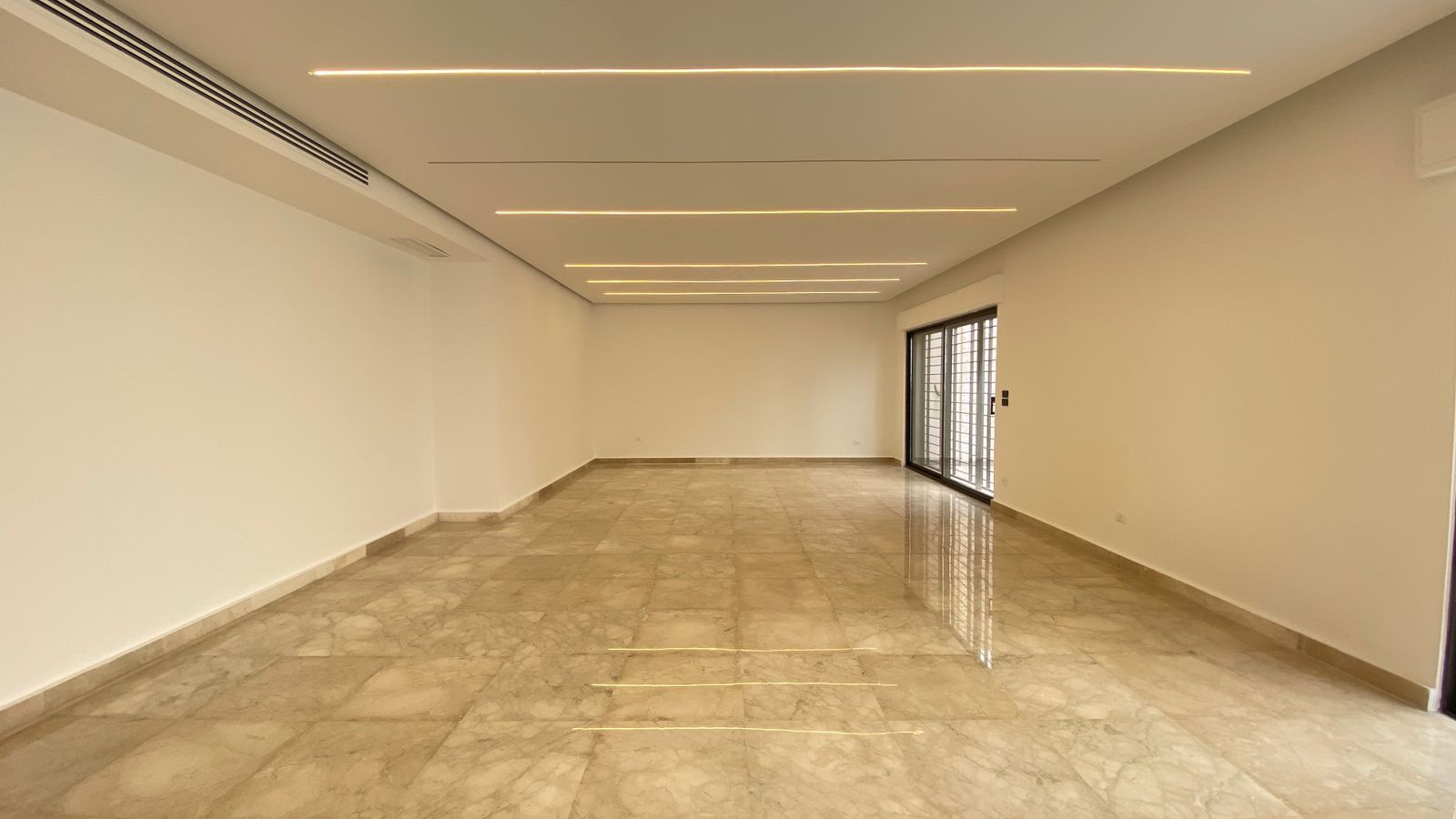 662e6bef-6b3b-4759-9474-0656d6f94fb4 Bright empty living space with cream marble floor, white walls, and slim recessed ceiling lights; sliding glass doors to a balcony on the right.