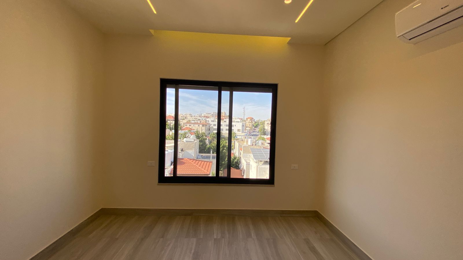 67321294-6a3d-466f-afe3-c021898eddff Empty rectangular room with beige walls and light wood flooring, featuring a large window overlooking a cityscape of buildings and red rooftops.