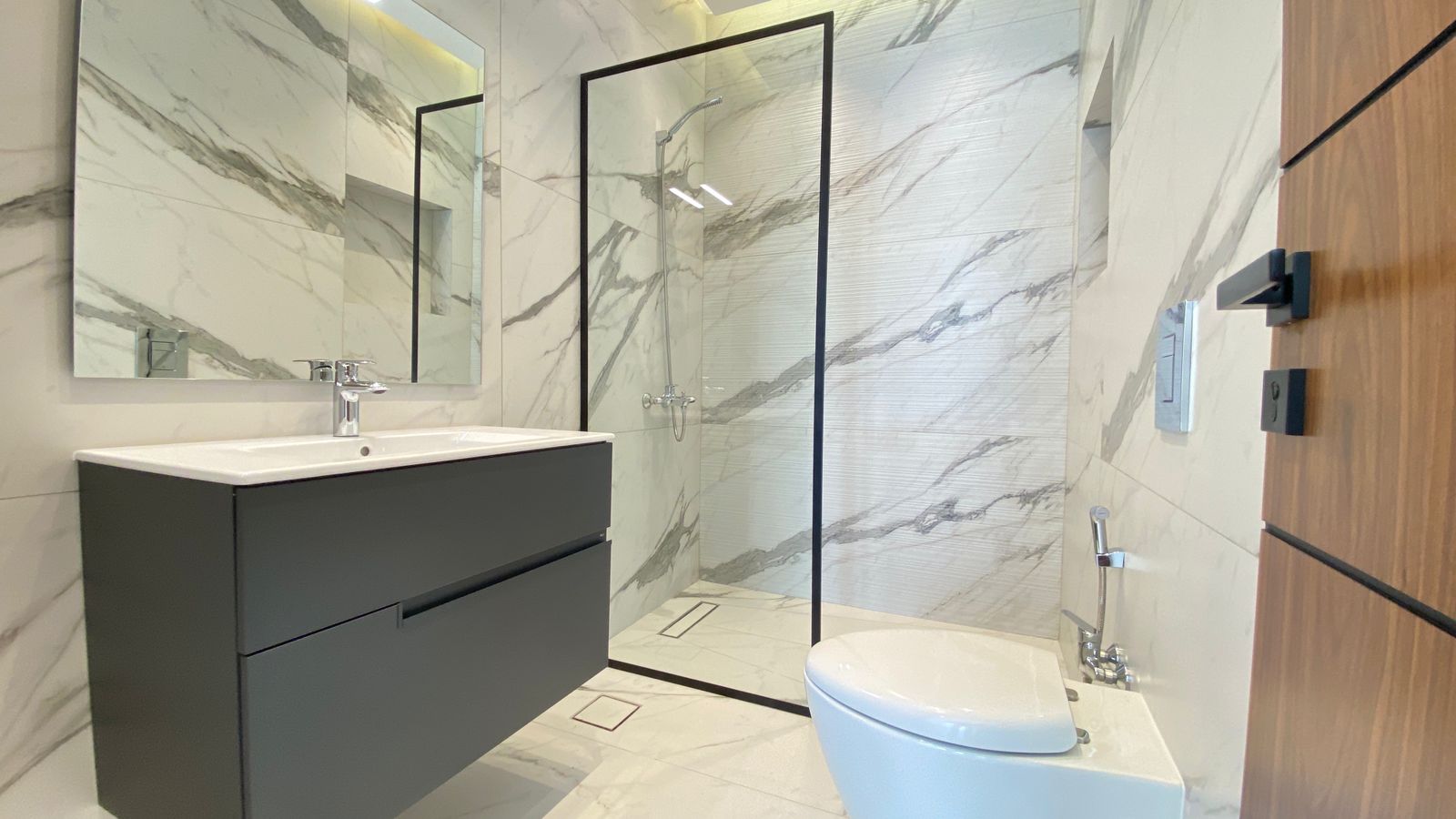 67bc7a1e-914e-4ad2-b9f1-6840d2ce48a5 Modern bathroom with marble walls, a dark floating vanity and white sink, and a glass-enclosed shower area nearby.