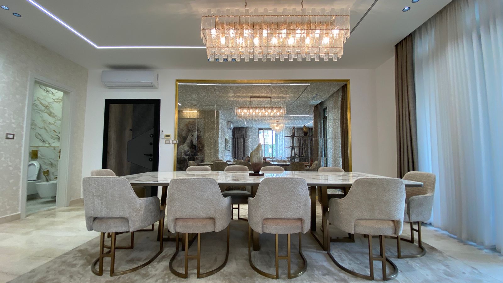 67bdc856-14bb-4ae8-9ec0-8f04c7fabbe1 Luxurious dining room with a long marble table, beige upholstered chairs, and a crystal chandelier above.
