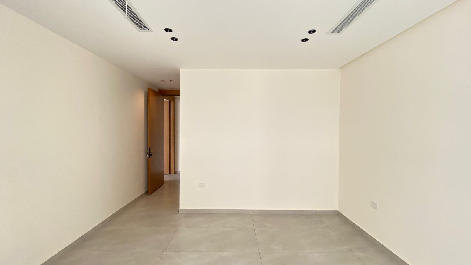 6813231e-93fa-49df-afca-d62442cd5d6c Empty rectangular room with light gray tile floor, white walls, and an open wooden door at the far end leading to a hallway.