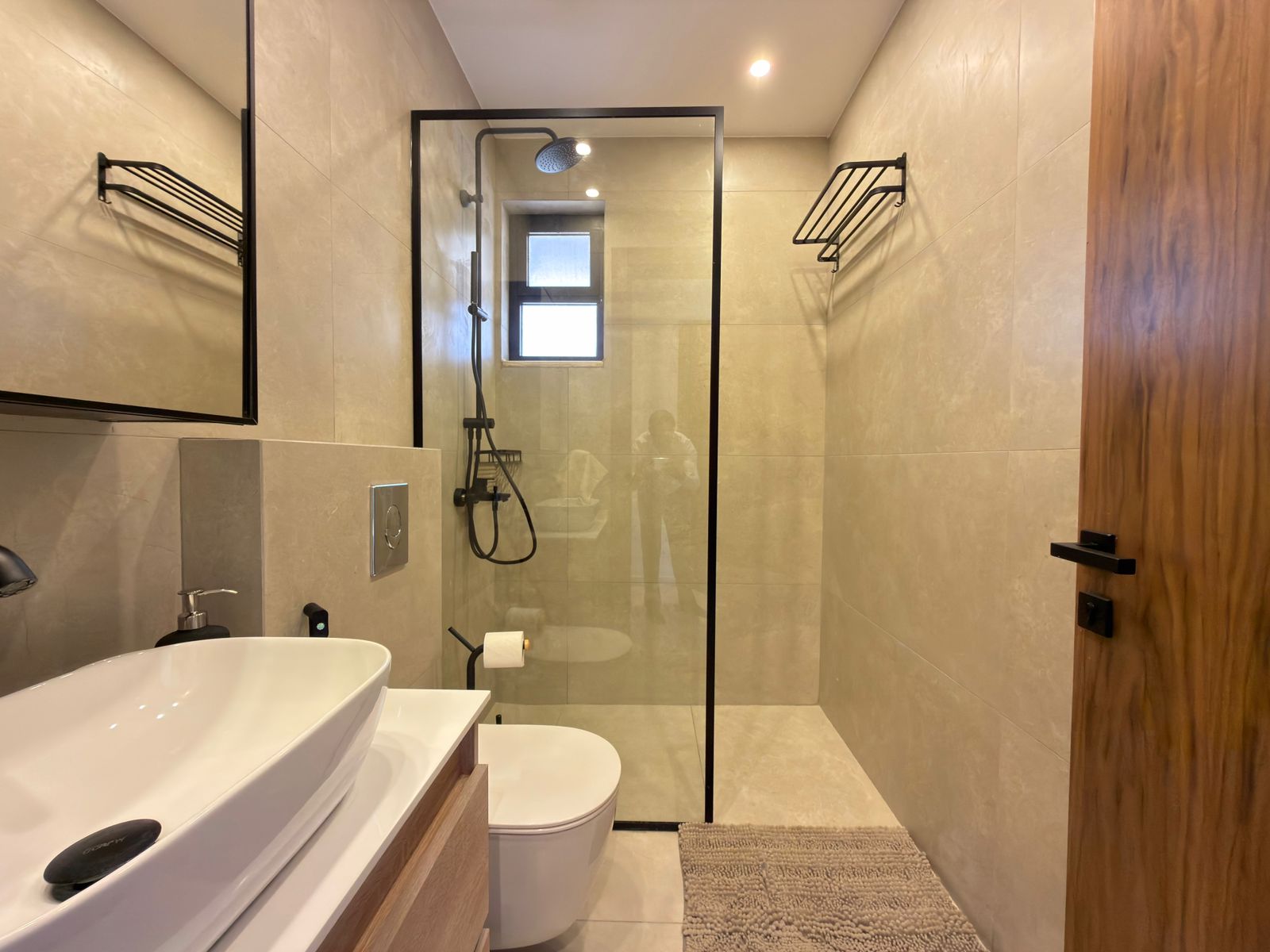 69a668d0-adfa-44bf-a6c6-1927525c10d0 Modern bathroom with a glass shower enclosure, beige tile walls, and a white freestanding sink on a wooden vanity.