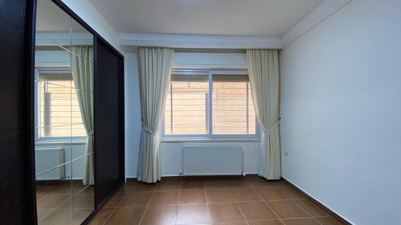 6a4dd54d-88c6-414b-a729-225d90c4e36f A bright, empty bedroom with large window, cream curtains, and a radiator beneath the window; brown tiled floor and a mirrored wardrobe on the left.