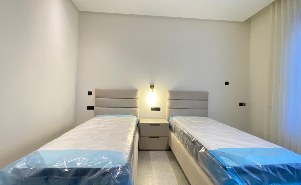 Twin bedroom with two single beds separated by a nightstand, padded headboards, and a wall lamp in a minimalist hotel room.