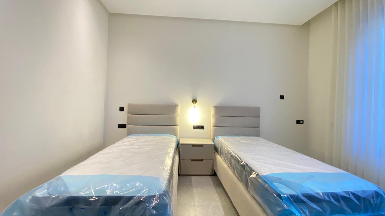 Twin bedroom with two single beds separated by a nightstand, padded headboards, and a wall lamp in a minimalist hotel room.