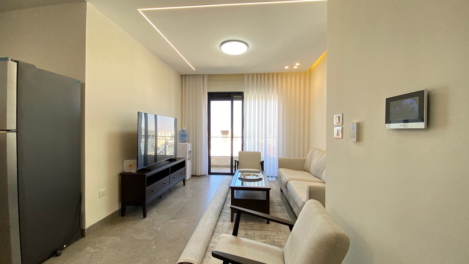 6b613eba-4729-40ad-8bc2-fa024f938b22 Living room with a beige sofa, glass coffee table, and a TV on a dark wooden stand facing a sliding door balcony with sheer white curtains.