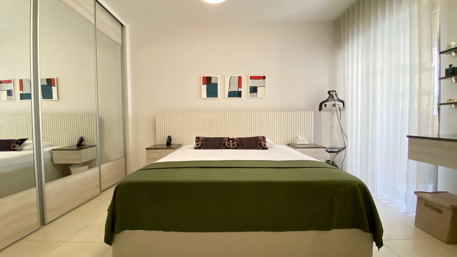 6bb044cc-bc91-4731-85af-549af89a0e63 Bright hotel-style bedroom with a white bed and olive-green cover, two nightstands, mirrored wardrobe, and abstract triptych above the headboard.