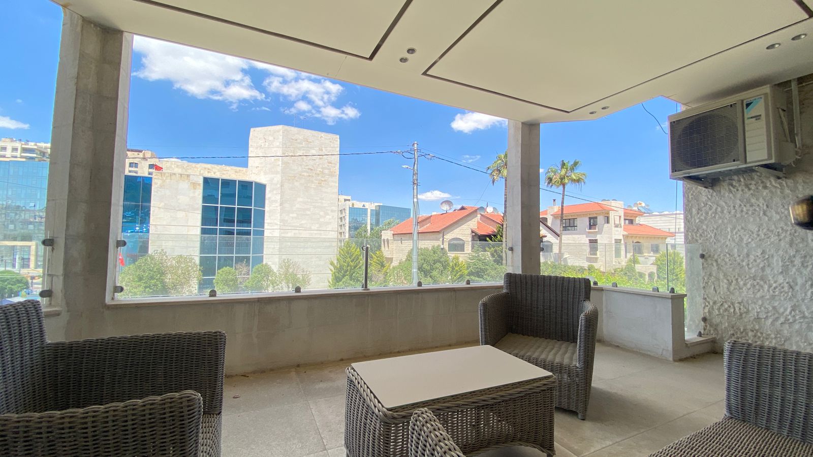 6bbfd35c-f7cf-4c21-8079-56ba0fcc3b4e Covered balcony with gray wicker seating and a rectangular coffee table, facing a glass railing and an urban street view under a blue sky.