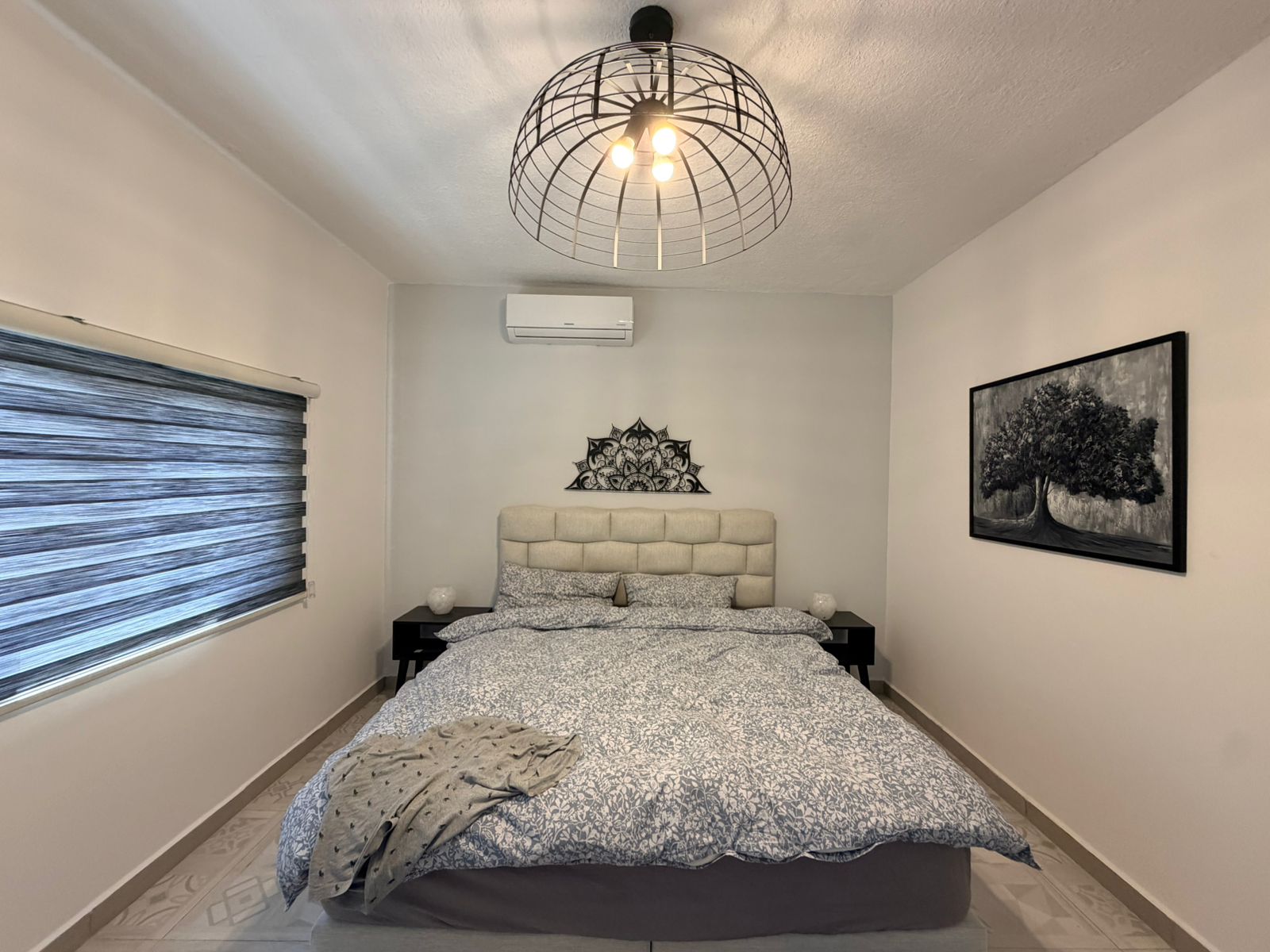 6ca2583b-b752-4dd6-82bb-66847a1085f9 Bedroom with an upholstered gray headboard, two black nightstands, and a decorative mandala above the bed; window with striped blinds on the left; ceiling cage light fixture; wall art of a tree on the right.