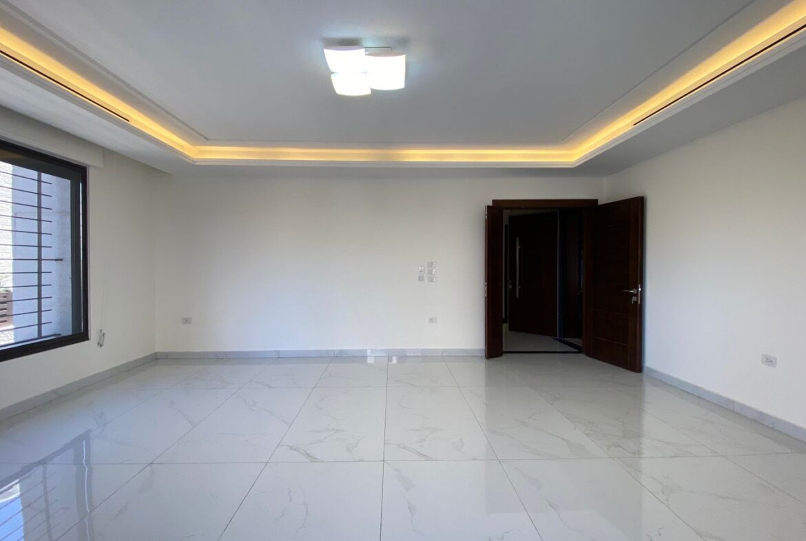 Empty living room with a large left-side window, white marble floor tiles, and dark wood double doors; warm recessed ceiling lighting.
