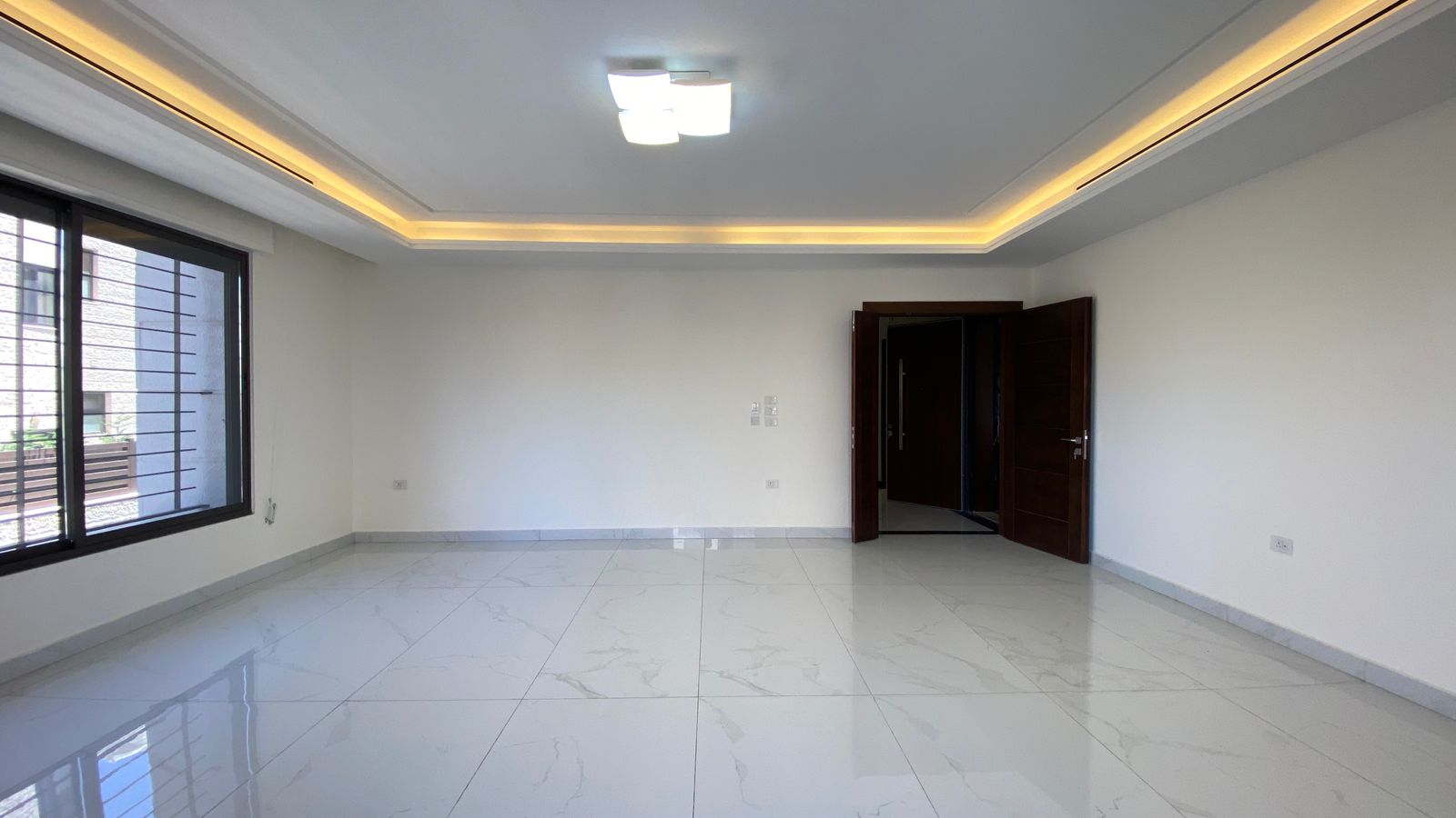 6cb6dd2e-3d29-4963-a70e-004bdb8f17ac Empty living room with a large left-side window, white marble floor tiles, and dark wood double doors; warm recessed ceiling lighting.