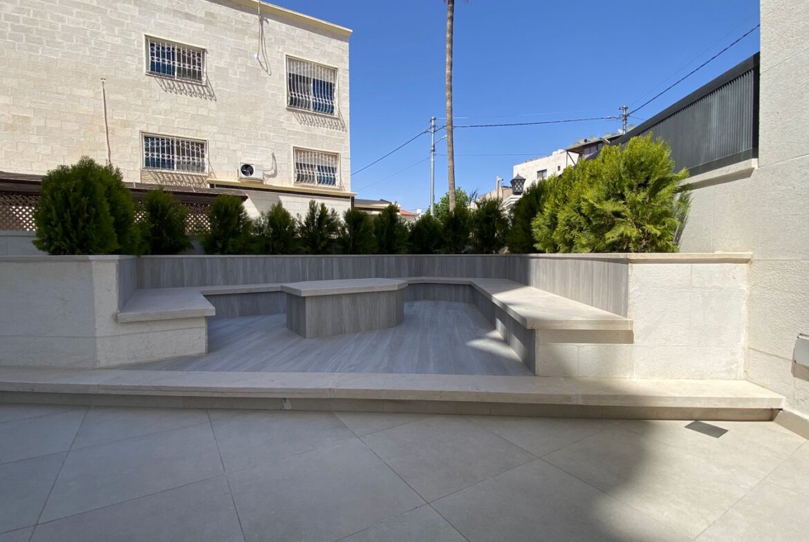 Stone-tiered outdoor patio with built-in benches and planters along a hedge under a clear blue sky