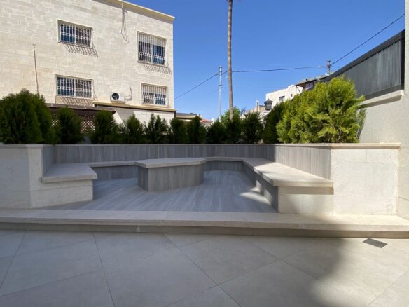 Stone-tiered outdoor patio with built-in benches and planters along a hedge under a clear blue sky