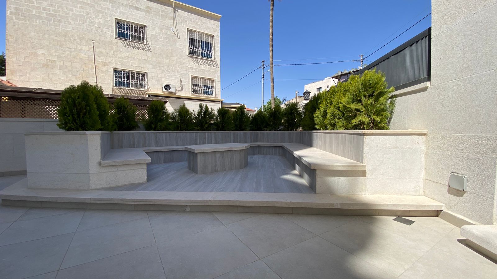 6d5b769b-c1d2-4253-9a35-7e605334f27a Stone-tiered outdoor patio with built-in benches and planters along a hedge under a clear blue sky