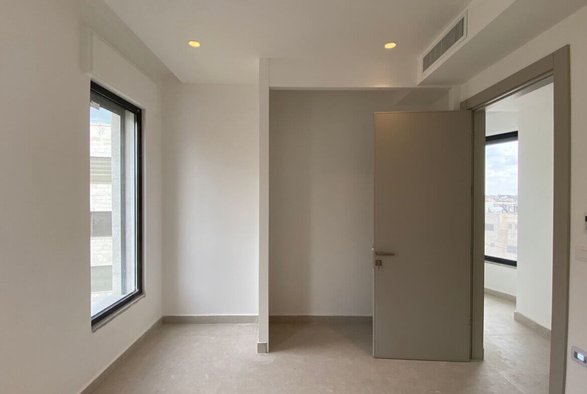 6dbb4ebe-9600-45ff-87d0-8e45bb191709 Empty modern room with a large window on the left and an open doorway to a brighter adjacent space with a city view.