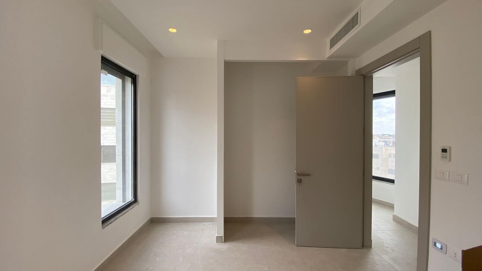 6dbb4ebe-9600-45ff-87d0-8e45bb191709 Empty modern room with a large window on the left and an open doorway to a brighter adjacent space with a city view.