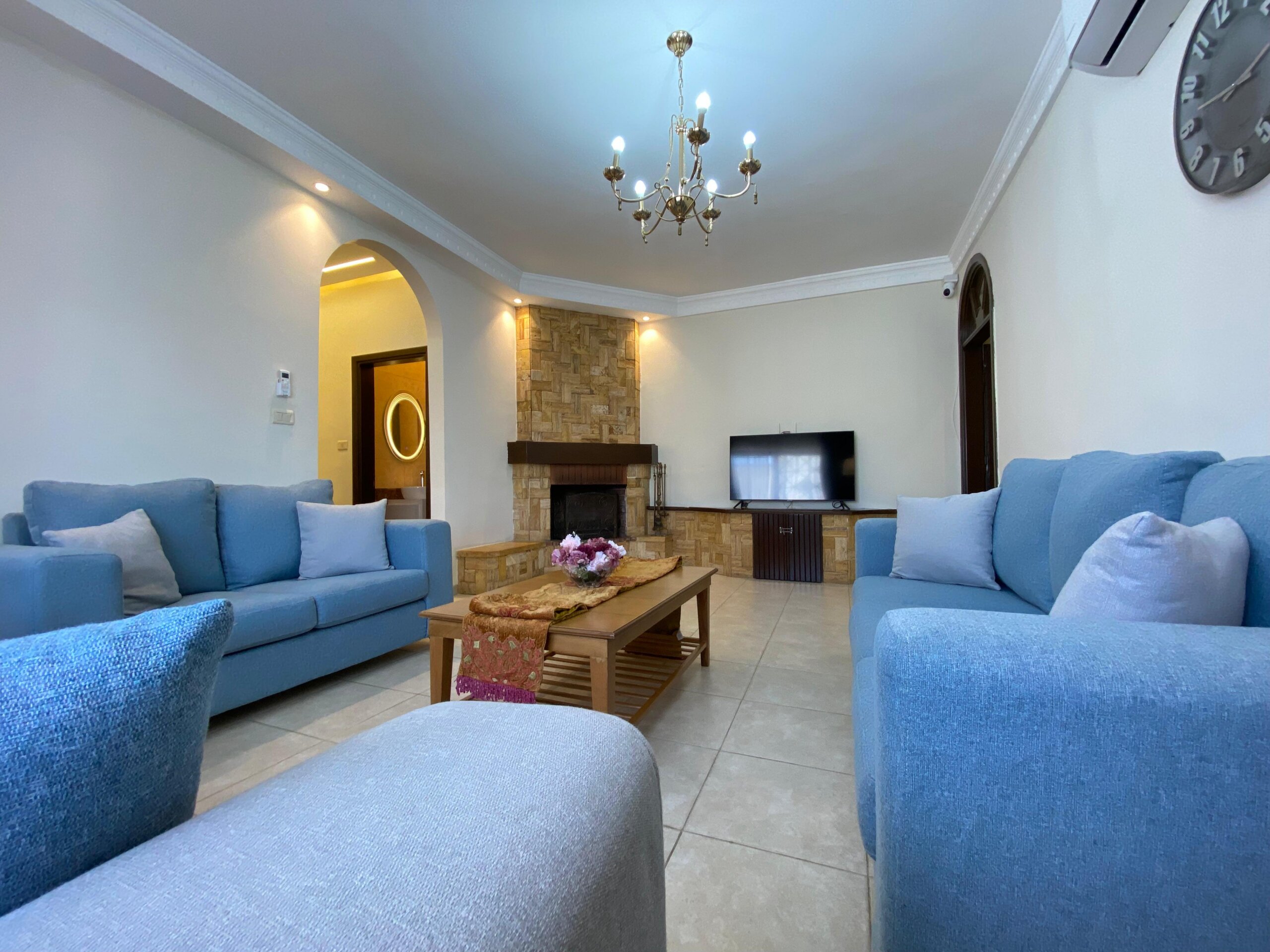 6e5707f1-6266-4ca7-b15b-5334e7d1ee51 Bright living room with light blue sofas arranged in a U-shape around a wooden coffee table and a stone fireplace centerpiece under a chandelier.