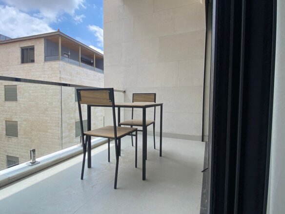 Balcony seating with a small wooden table and two matching chairs overlooking a cityscape, glass railing in the foreground.