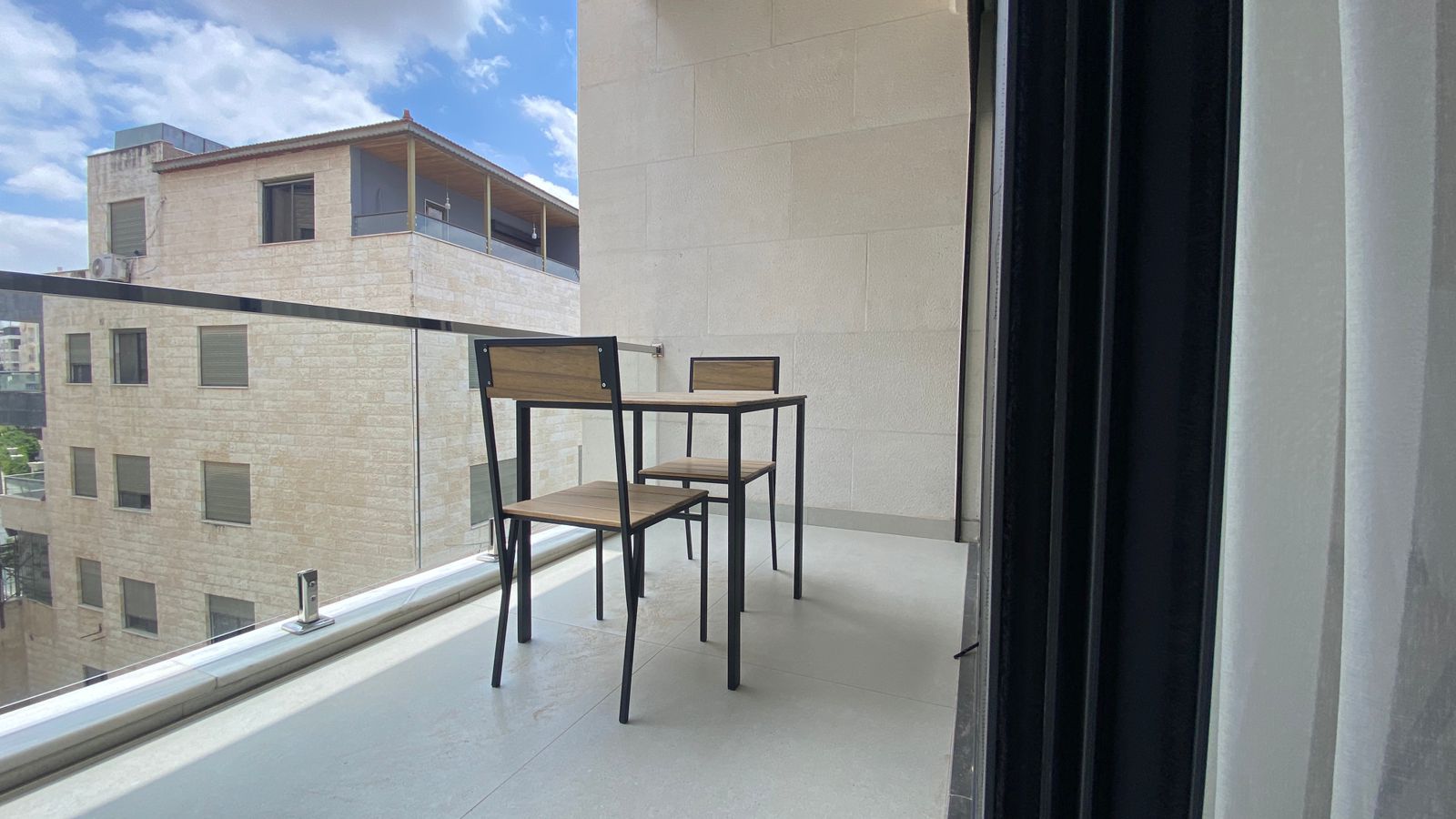 6ecc8314-0039-4dd7-afa0-2fde6acf1c24 Balcony seating with a small wooden table and two matching chairs overlooking a cityscape, glass railing in the foreground.