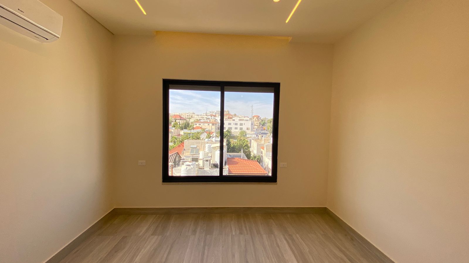 6ee5b50e-d4d8-4ce8-af4d-e045d087e0fc Empty beige room with a large window showing a cityscape; wall-mounted air conditioner on the left and light wood flooring.