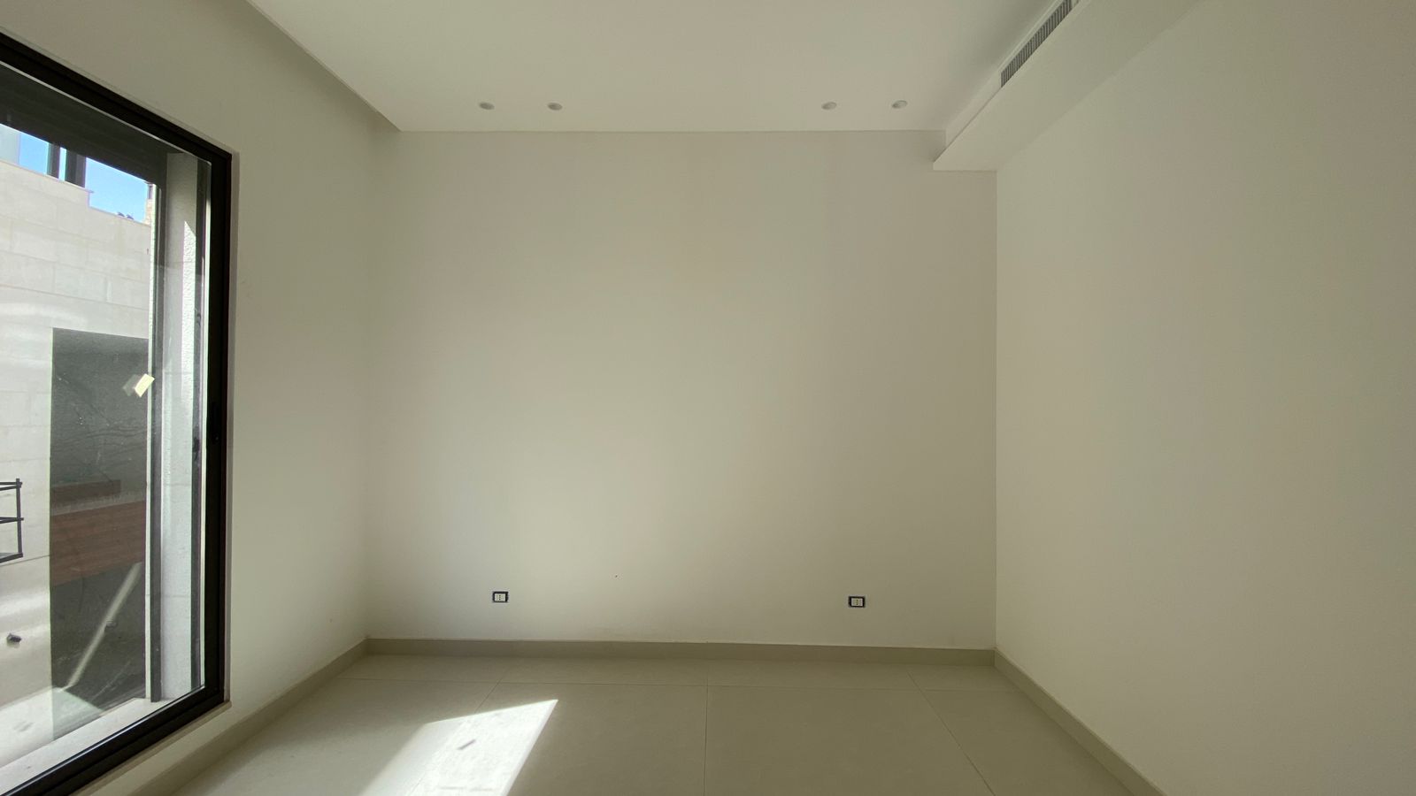 6ef4165d-984f-46cf-bc97-df9875468613 Empty white room with a large glass window on the left, sunlight on the tiled floor, and wall outlets nearby.
