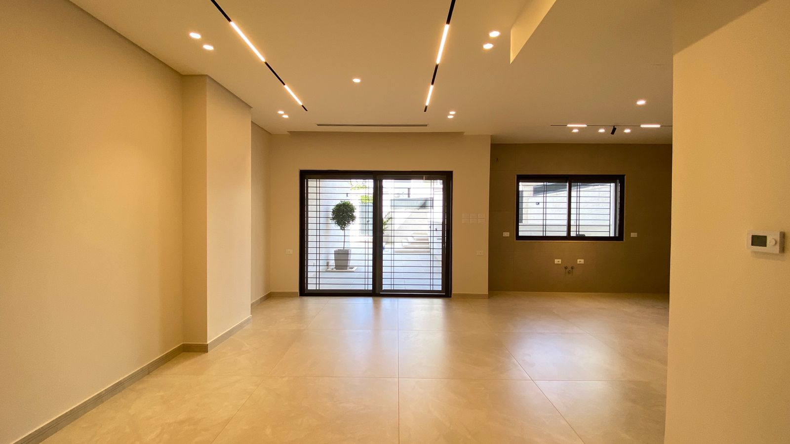 6fa5b243-e3d5-4ea1-8639-e5d0163e4a7e Empty contemporary living space with beige walls, large sliding glass doors to a patio and a potted tree outside; warm ceiling lights.