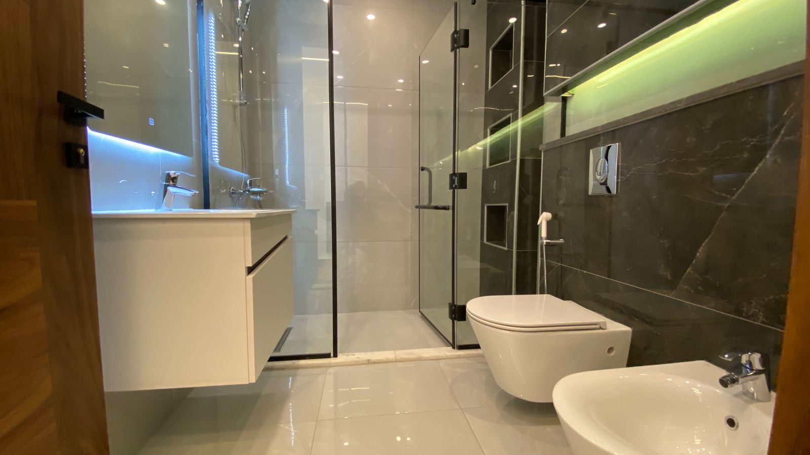 6febf2c3-0ce7-4e7c-a84f-3c9d2f8549bc Modern bathroom with a glass-enclosed shower, wall-hung white vanity, and wall-mounted toilet and bidet, set against dark marble with green lighting accents.