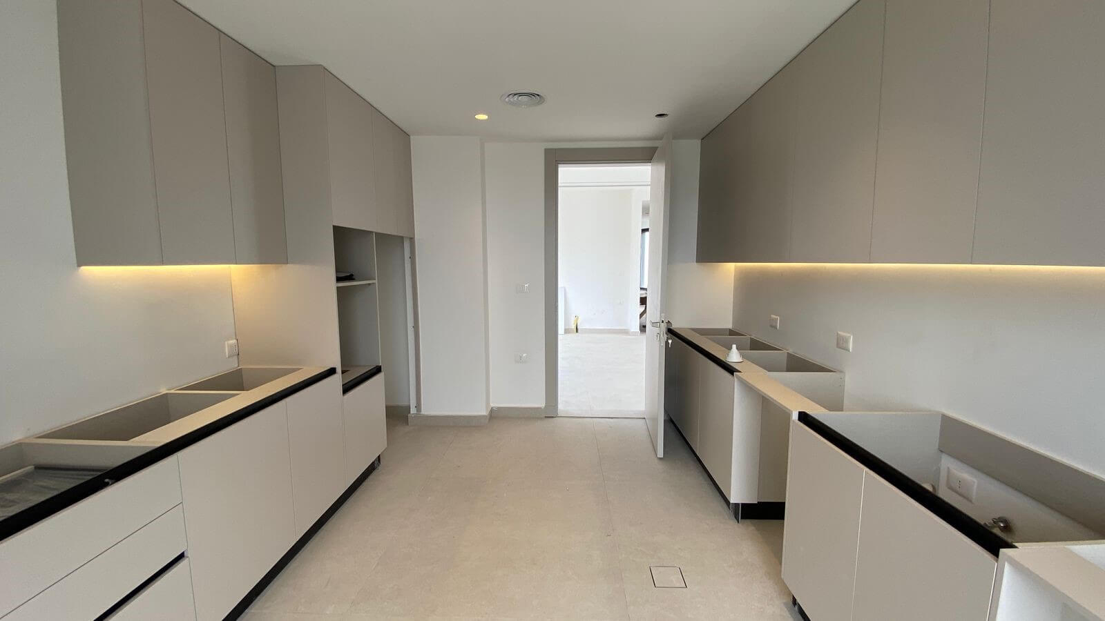 6ff52f97-4725-4216-beb9-a7c5c2ac401f Galley-style modern kitchen with light gray flat-panel cabinets, white walls, and warm under-cabinet lighting along the backsplash.