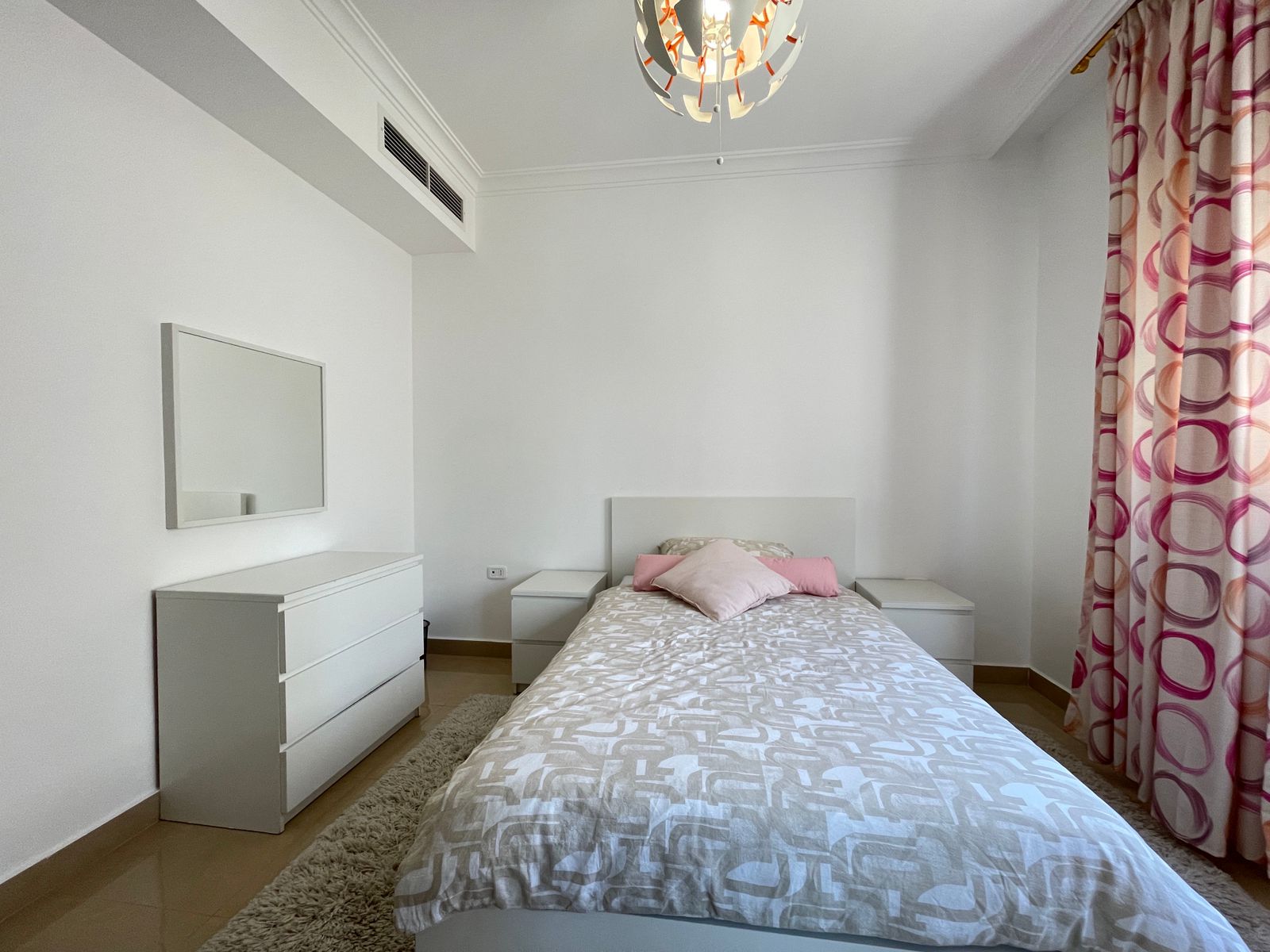 7099c269-1ae2-47d7-ad57-0d58ebadca09 Minimalist white bedroom with a low bed, light gray patterned bedding, and pink pillows; white dressers, a wall mirror, and pink circular curtains on the right.