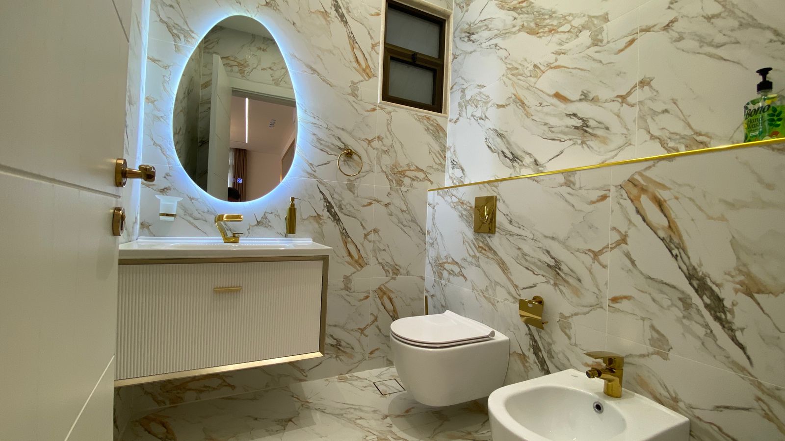 709a54ae-5f41-4cdc-b17b-b56738e66e0e Luxurious marble bathroom with gold fixtures and an oval backlit mirror above a white vanity.