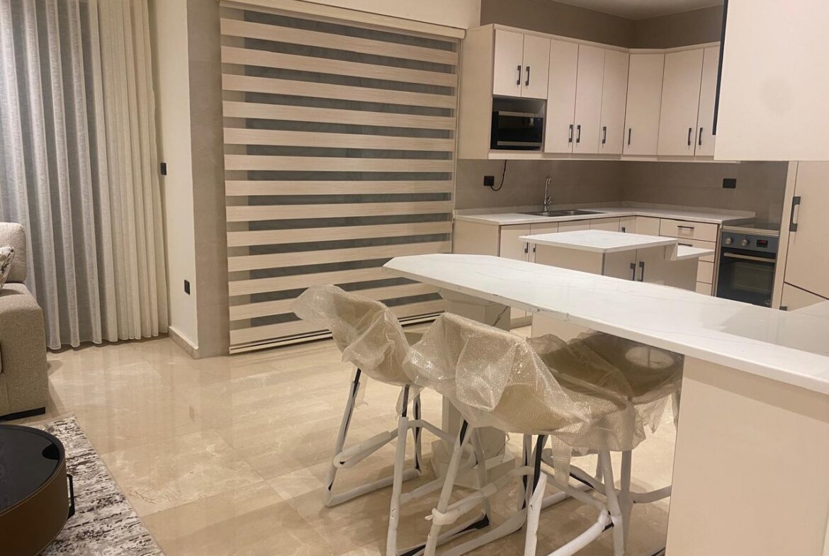 Modern white kitchen with a long island and three plastic-wrapped stools; built-in oven, microwave, and white cabinets with striped window blinds in the background.