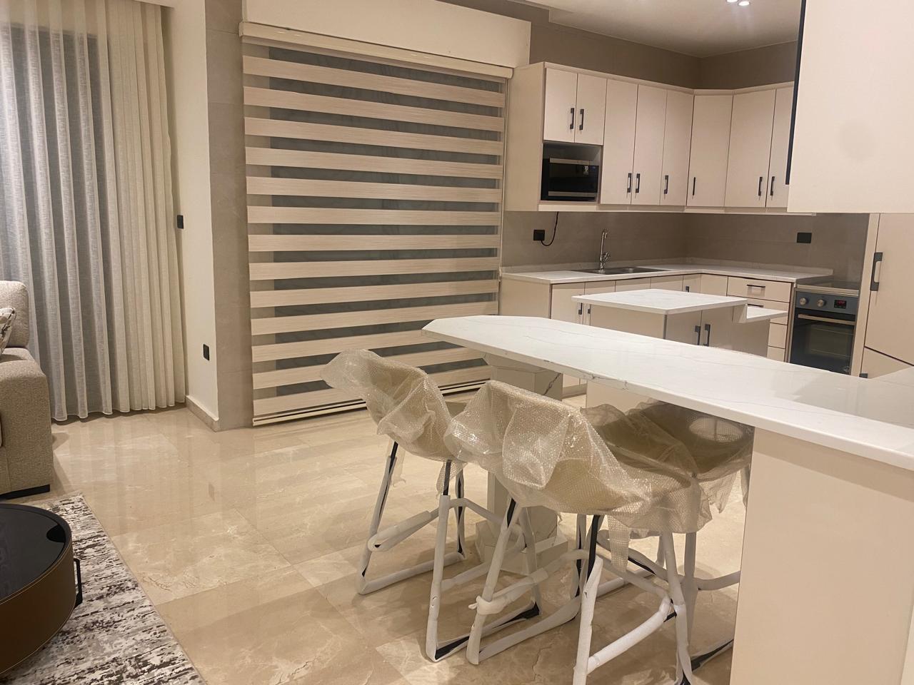 Modern white kitchen with a long island and three plastic-wrapped stools; built-in oven, microwave, and white cabinets with striped window blinds in the background.