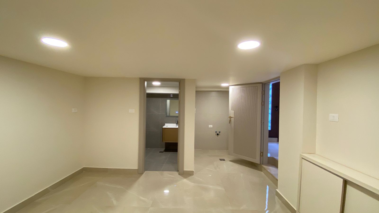 713045db-8c72-4d45-aa3a-0c4860c7ee0a Basement with beige walls and a glossy tile floor, two doorways; left leads to a bathroom with a vanity visible through the door.