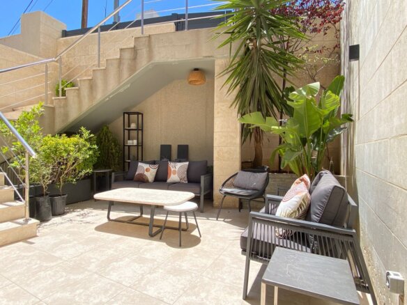 Modern outdoor patio with gray seating, coffee table, and potted plants under a shaded area near a stairway.