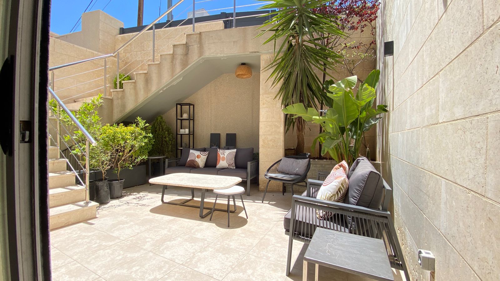 72b8ad04-fac9-4939-a005-86478e2432b4 Modern outdoor patio with gray seating, coffee table, and potted plants under a shaded area near a stairway.