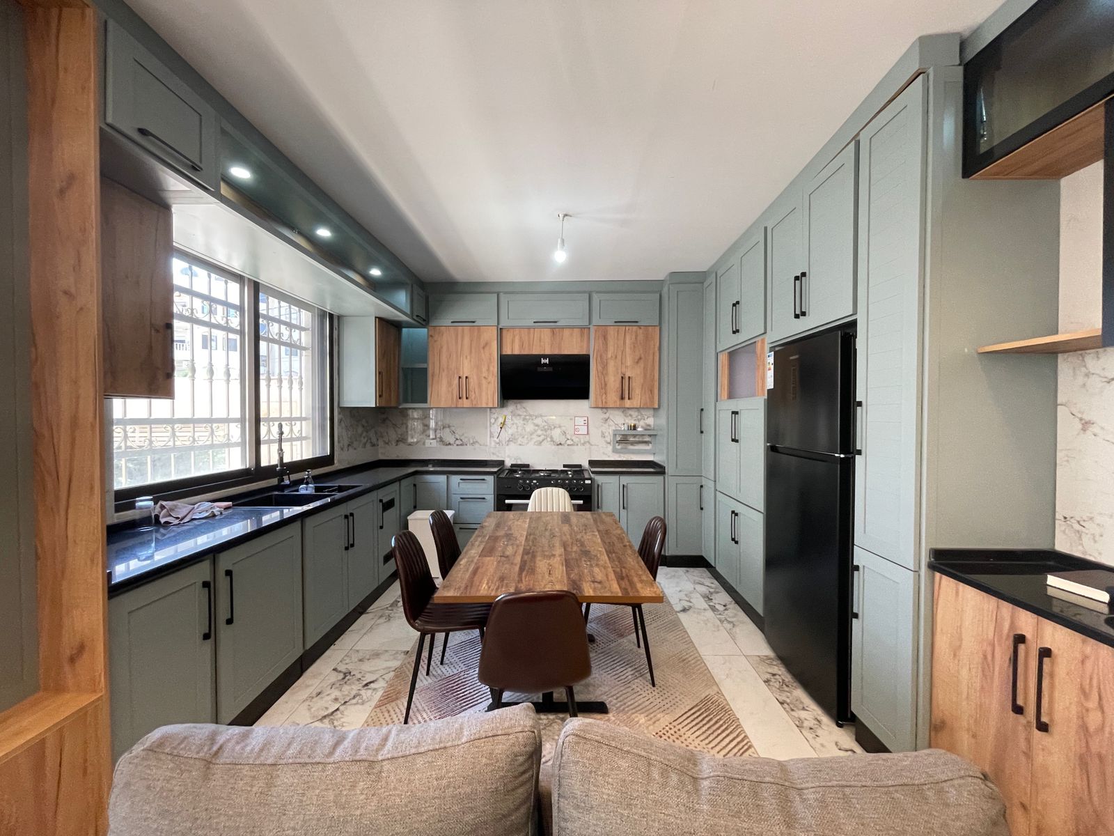 739997ed-b863-4927-86a3-4229f7365c59 Modern kitchen with sage green cabinets, marble backsplash, and a wooden dining table in the center.