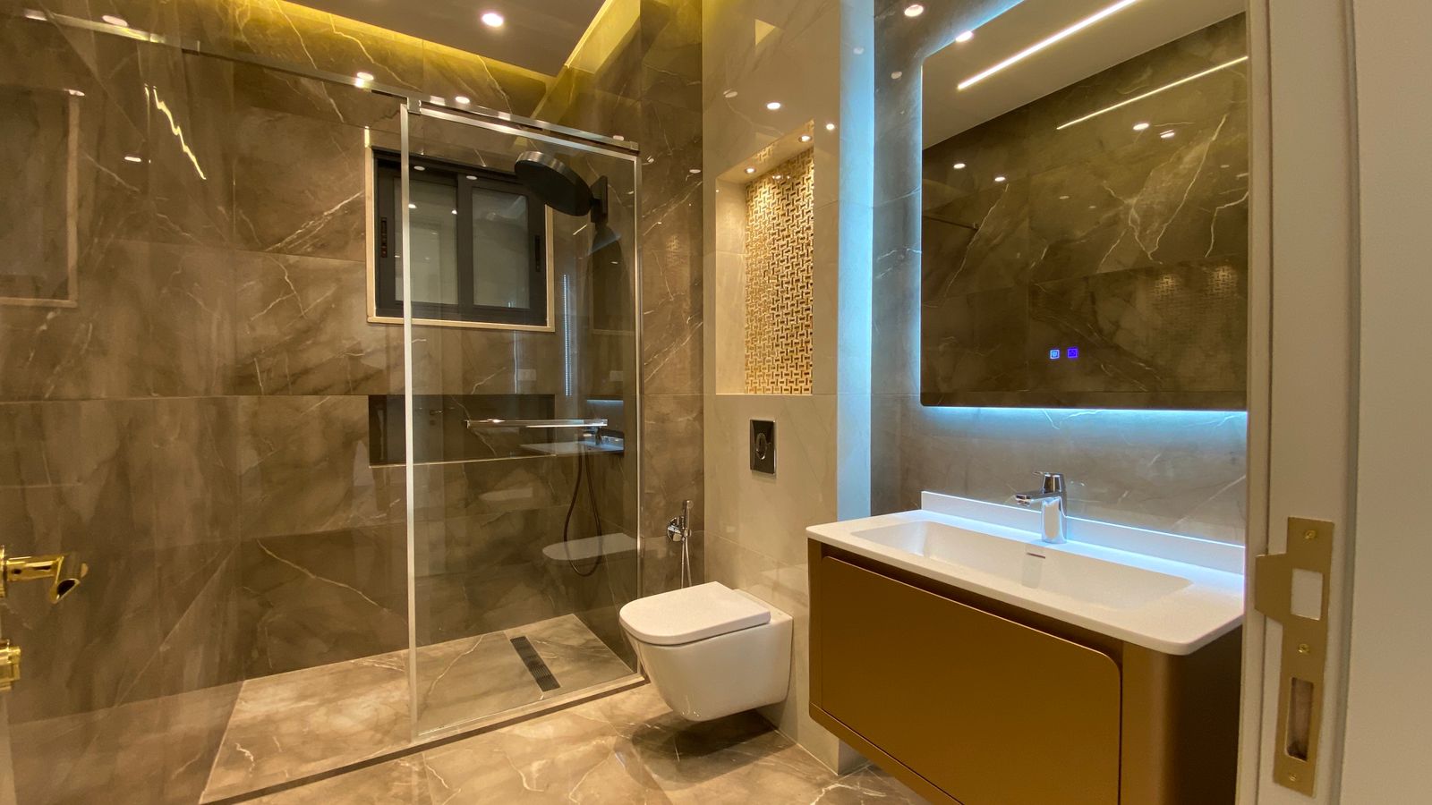 73d7ffbd-8ee1-41b8-b6c6-d5461c51c475 Modern marble bathroom with a glass-enclosed shower, wall-mounted toilet, and a brown vanity with a backlit mirror.