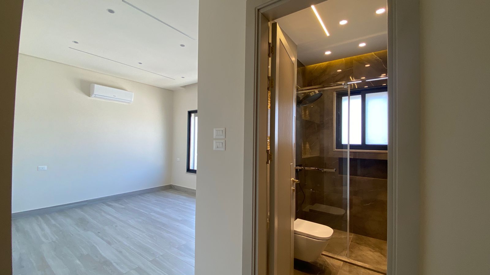 7407ef8b-08d8-43ad-a638-ee3f15020fd2 Empty living room with light walls, gray wood flooring, and a wall-mounted AC; doorway to a glass-shower bathroom.
