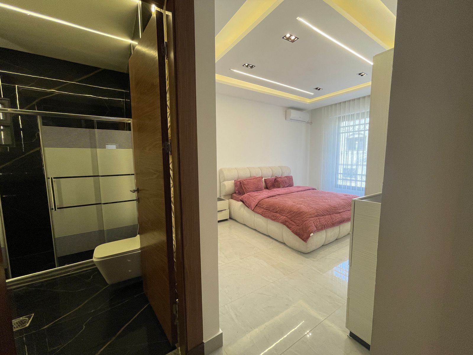 7448f77d-0fd7-4aa7-b5af-a25f7c4ef1f9 Modern bedroom with a tufted cream bed, pink bedding, white nightstands, and sheer-curtained window; doorway to a black tiled bathroom on the left.