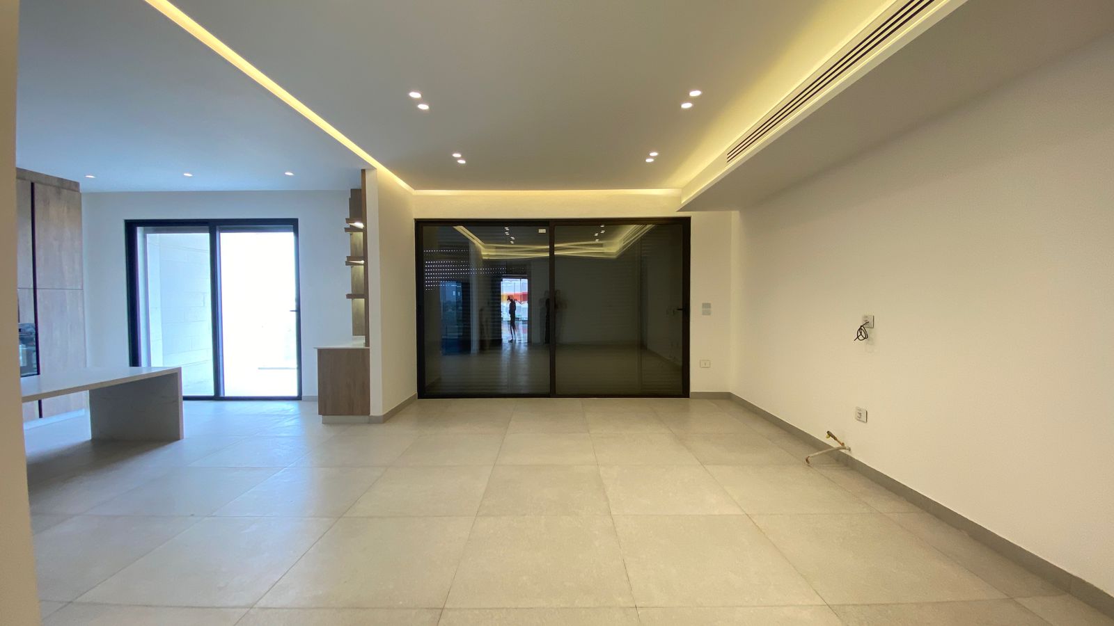 75475a24-736b-4740-bc3a-ea689ca99980 Empty, modern open-plan living area with light tile floor, white walls, and a large dark-framed glass door at the far end.