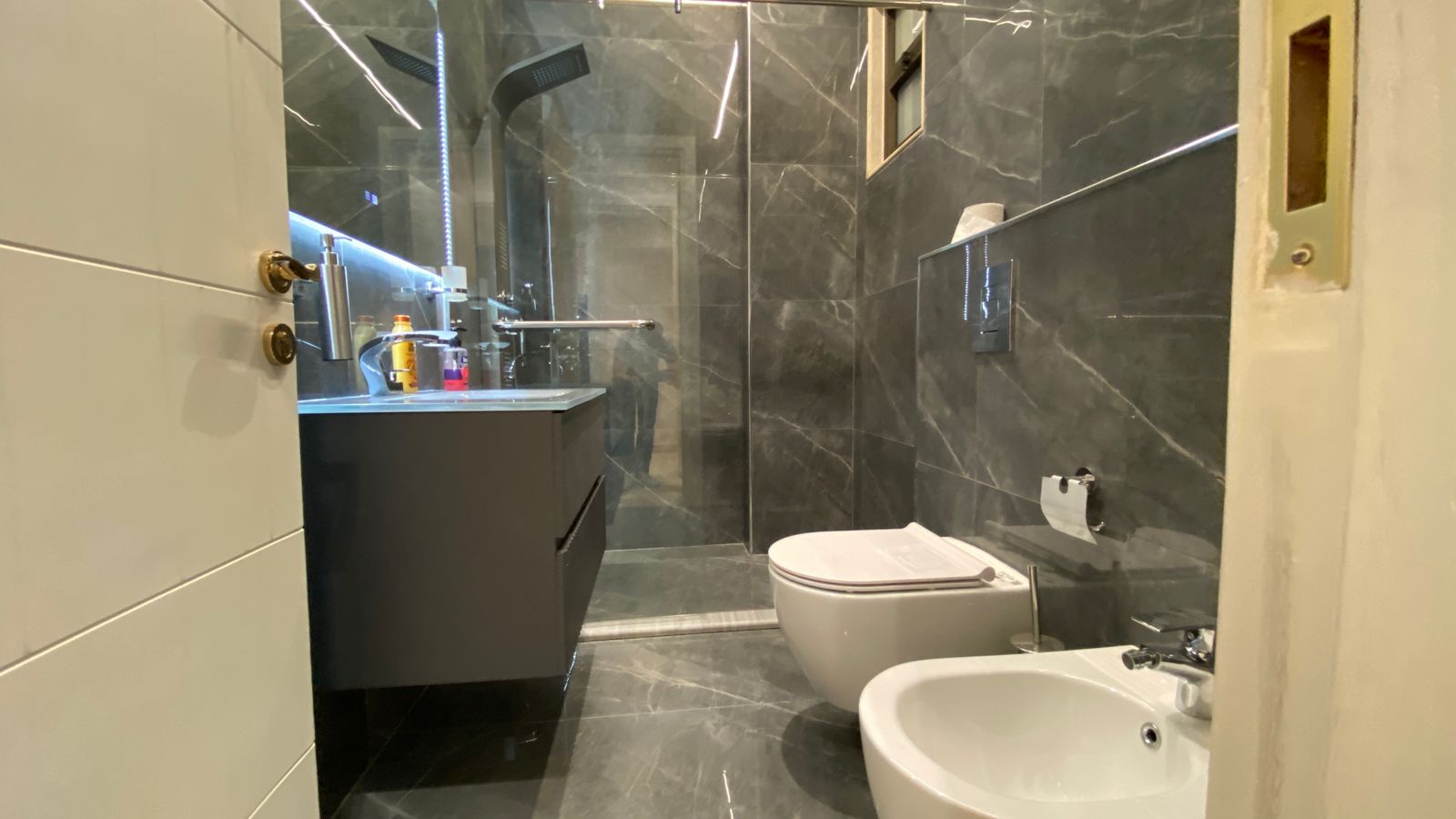 75b2f637-8544-40e3-be6d-388359e25824 Modern marble bathroom with glass shower, floating vanity, wall-mounted toilet and bidet. A few toiletries sit on the sink.