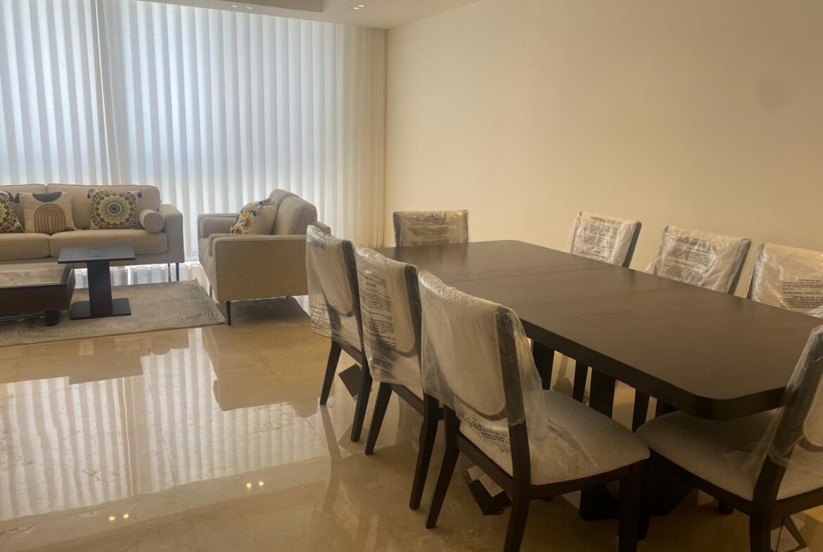 Open-plan living and dining area with a dark wood dining table and chairs covered in plastic, beige sofa set, and vertical blinds.