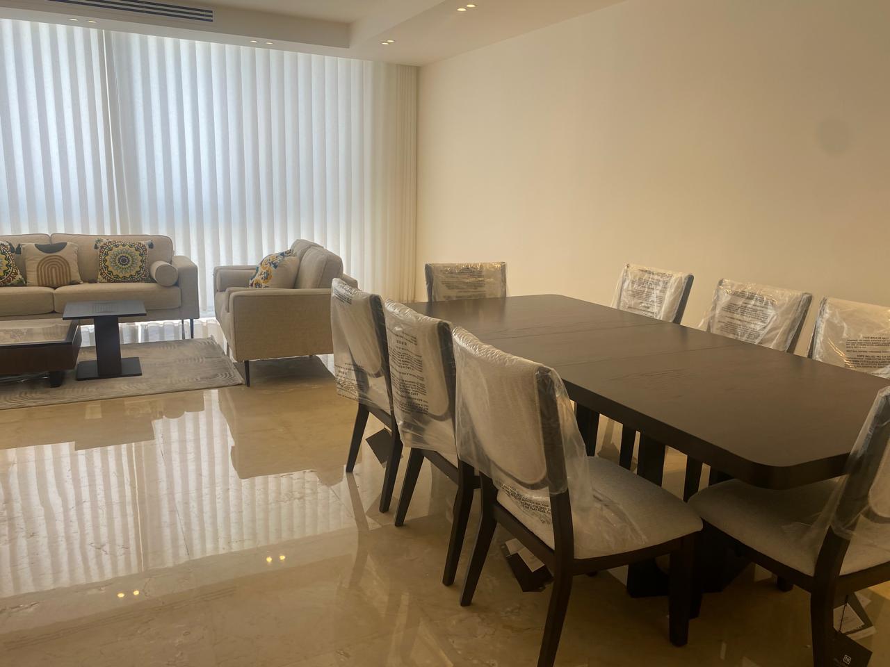 Open-plan living and dining area with a dark wood dining table and chairs covered in plastic, beige sofa set, and vertical blinds.