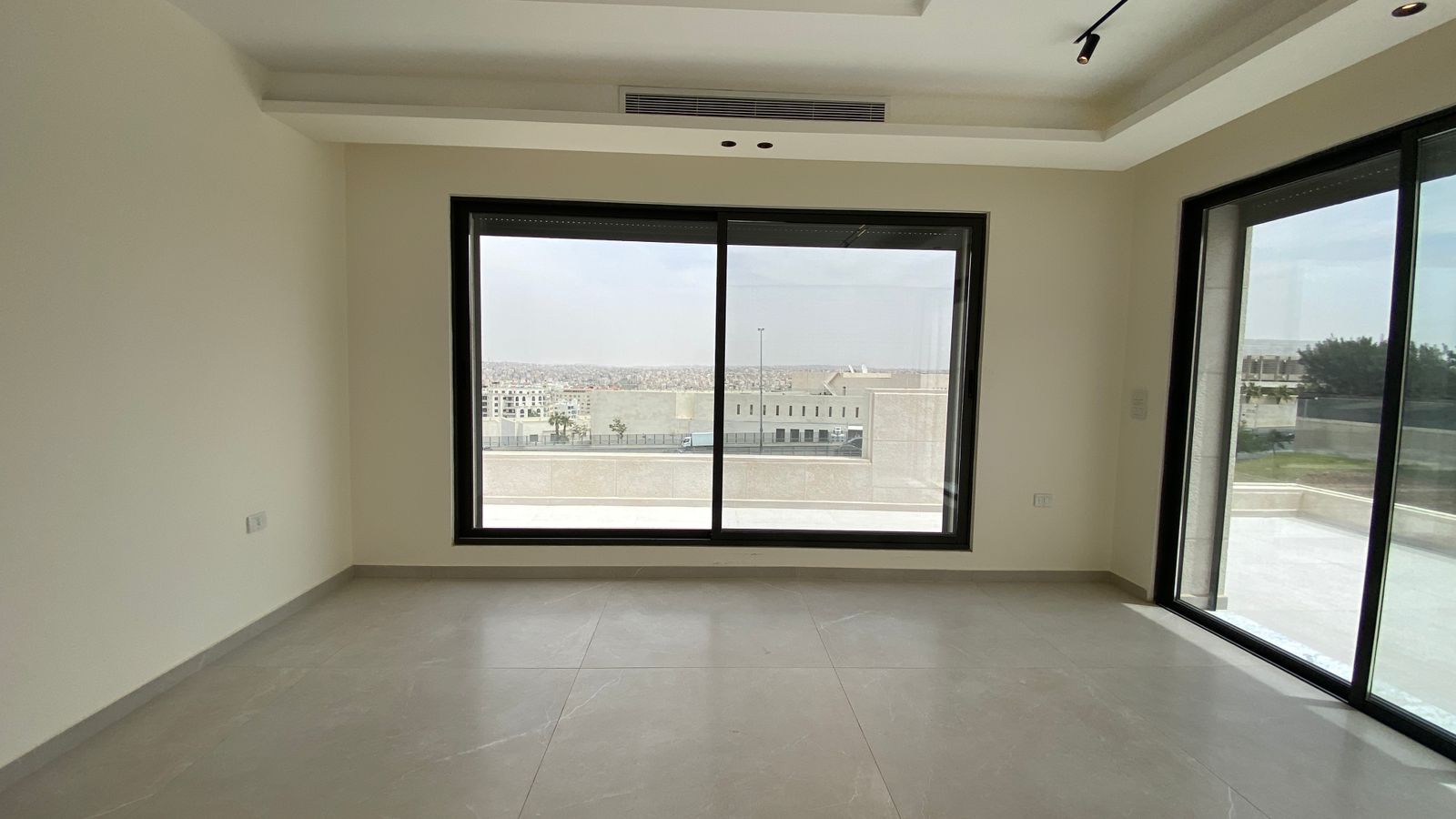 76e4b7dc-e3d3-4098-9958-743aec26fbab Empty modern living room with beige walls, large sliding window and a glass door to a balcony, overlooking a cityscape.