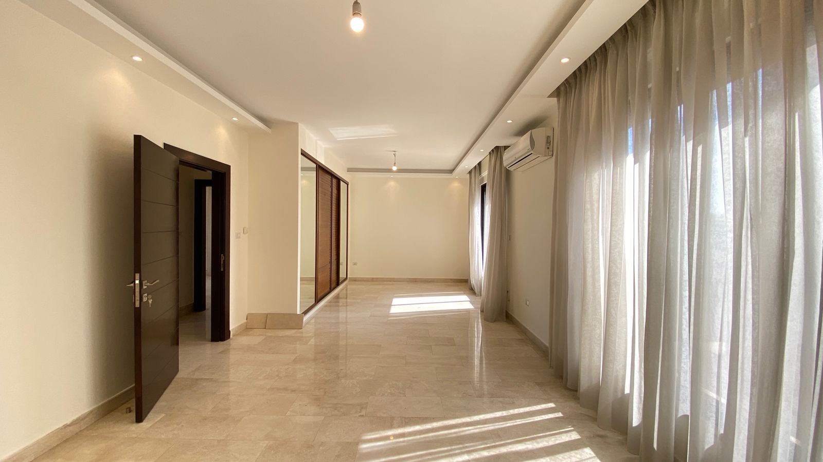 774182a8-6177-4cec-be59-3431ad0027ad Bright empty living room with beige marble floor, large windows with sheer curtains, and a wall-mounted air conditioner.